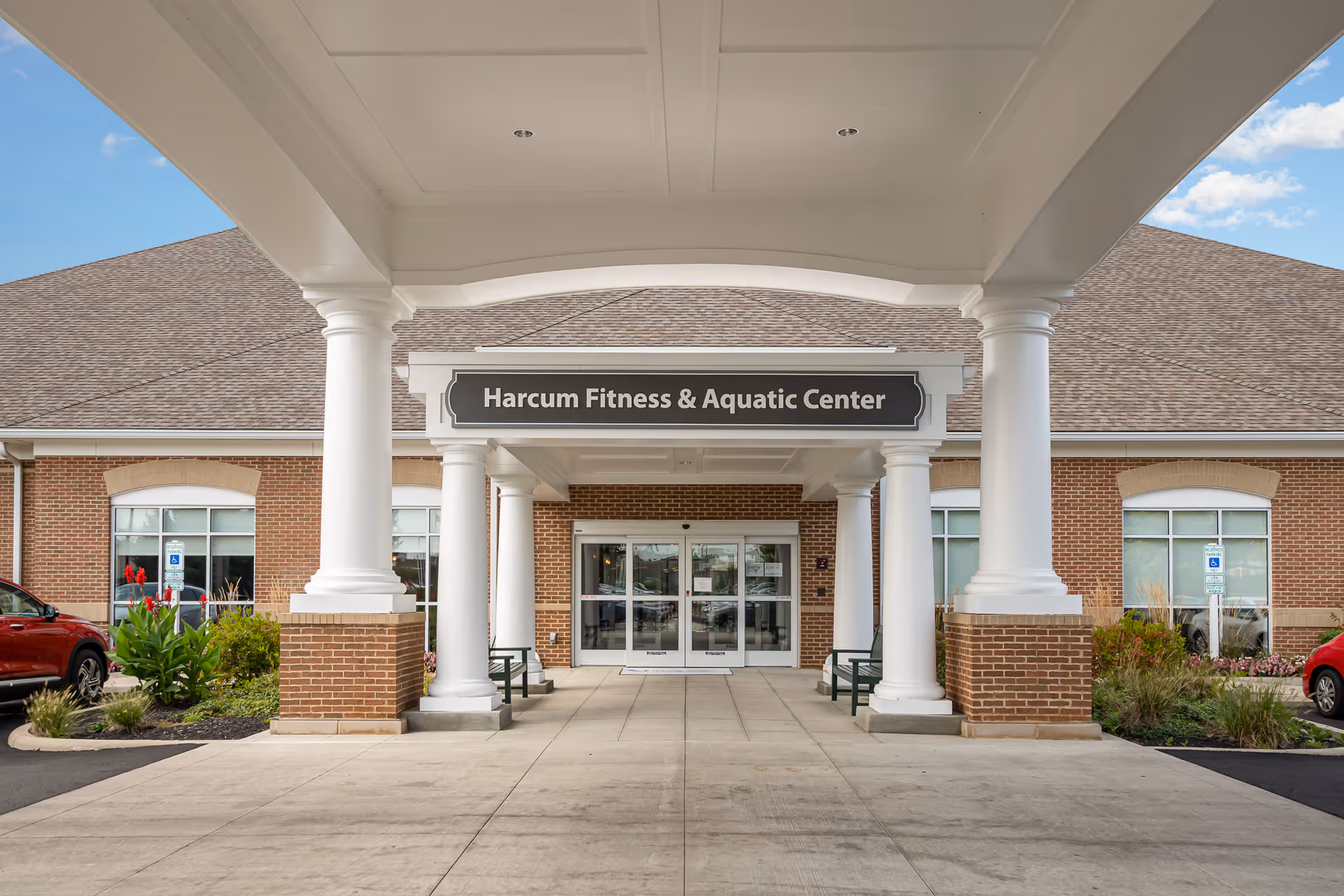 Entrance to the Harcum Fitness & Aquatic Center at Wesley Ridge, featuring a covered walkway with white columns, brick walls, glass double doors, and a sign above the entrance. There are plants and parked cars visible on either side.