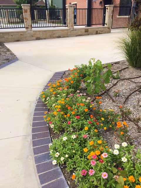 A landscaped outdoor area with a curved concrete walkway bordered by a flower bed filled with colorful flowers including orange, pink, white, and yellow blooms. In the background, there is a stone and metal fence enclosing the area.