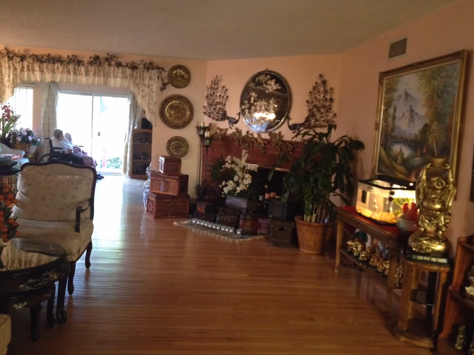 A cozy living room with wooden flooring, decorated with various plants, ornate wall hangings, and a fireplace adorned with flowers and candles. There is a large painting on the right wall above a table holding a fish tank and a golden statue. In the background, two elderly people are seated near a sliding glass door with lace curtains.