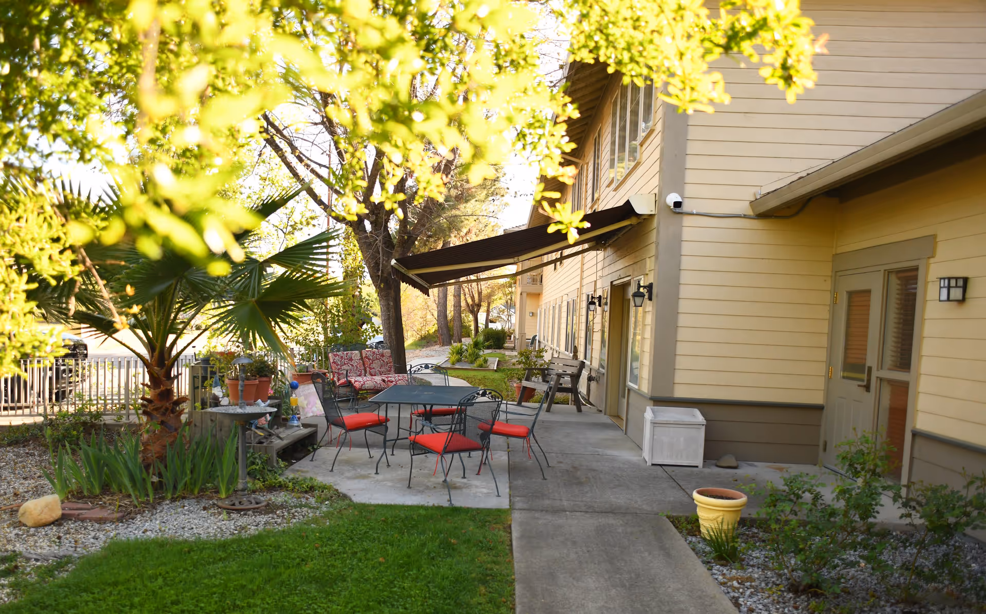 Outdoor patio area at Oakdale Heights Assisted Living featuring a concrete seating area with a black metal table and chairs with red cushions, a bench with floral cushions, potted plants, a small palm tree, and a beige building with multiple windows and doors in the background.