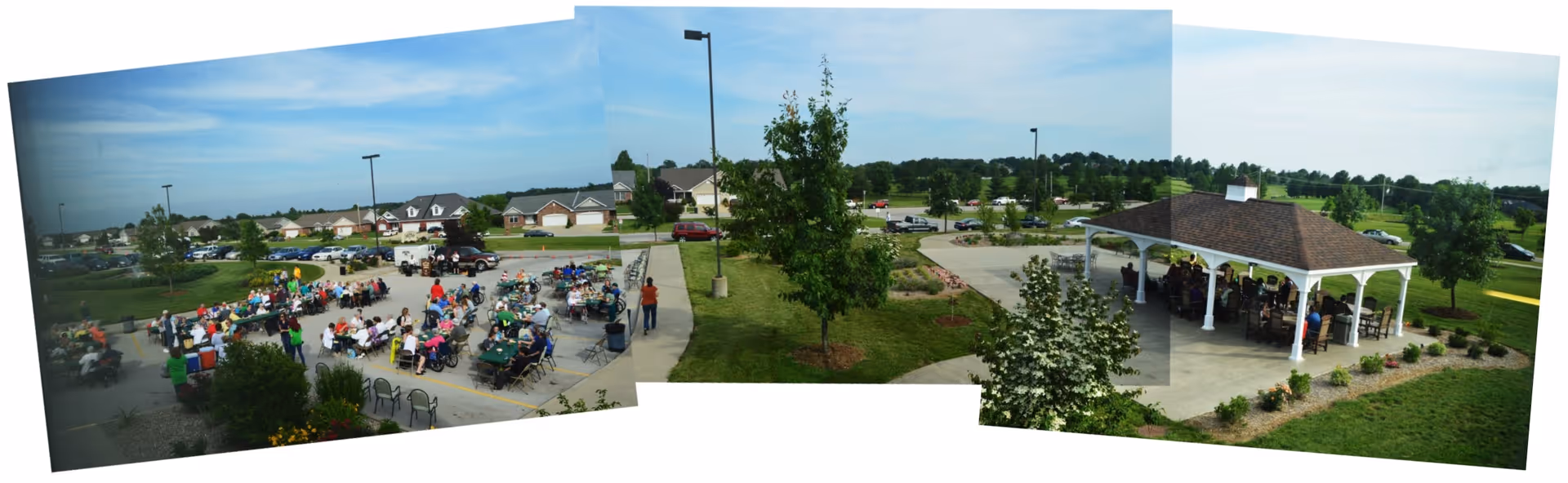 Outdoor gathering at Magnolia Terrace Senior Living Apartments with many people seated at tables in a parking lot area on the left, and a covered pavilion with more people seated on the right, surrounded by green lawns, trees, and residential houses in the background under a blue sky.