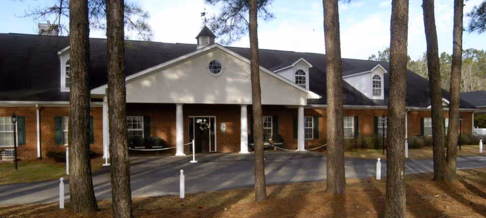 Front entrance of a single-story brick senior living building with white columns and pine trees in the foreground.