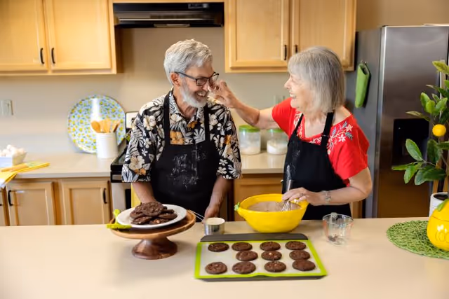 An elderly man and woman wearing aprons are in a kitchen baking cookies. The woman is playfully feeding the man a cookie while smiling. There are freshly baked cookies on a tray and a cake stand on the counter, along with a yellow mixing bowl and kitchen utensils in the background.