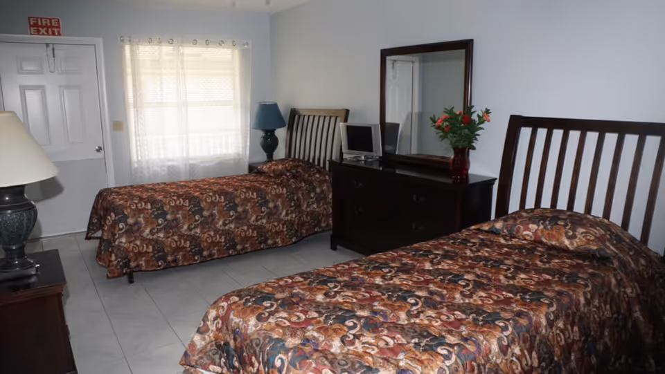 A bedroom with two twin beds covered in patterned brown and orange bedspreads. Between the beds is a dark wooden dresser with a large mirror, a small TV, and a vase with red flowers. There are two lamps, one on a nightstand near the bed in the foreground and another on a table near the bed by the window. The room has white tiled floors, white walls, and a window with sheer white curtains. A door with a red 'FIRE EXIT' sign above it is visible in the background.