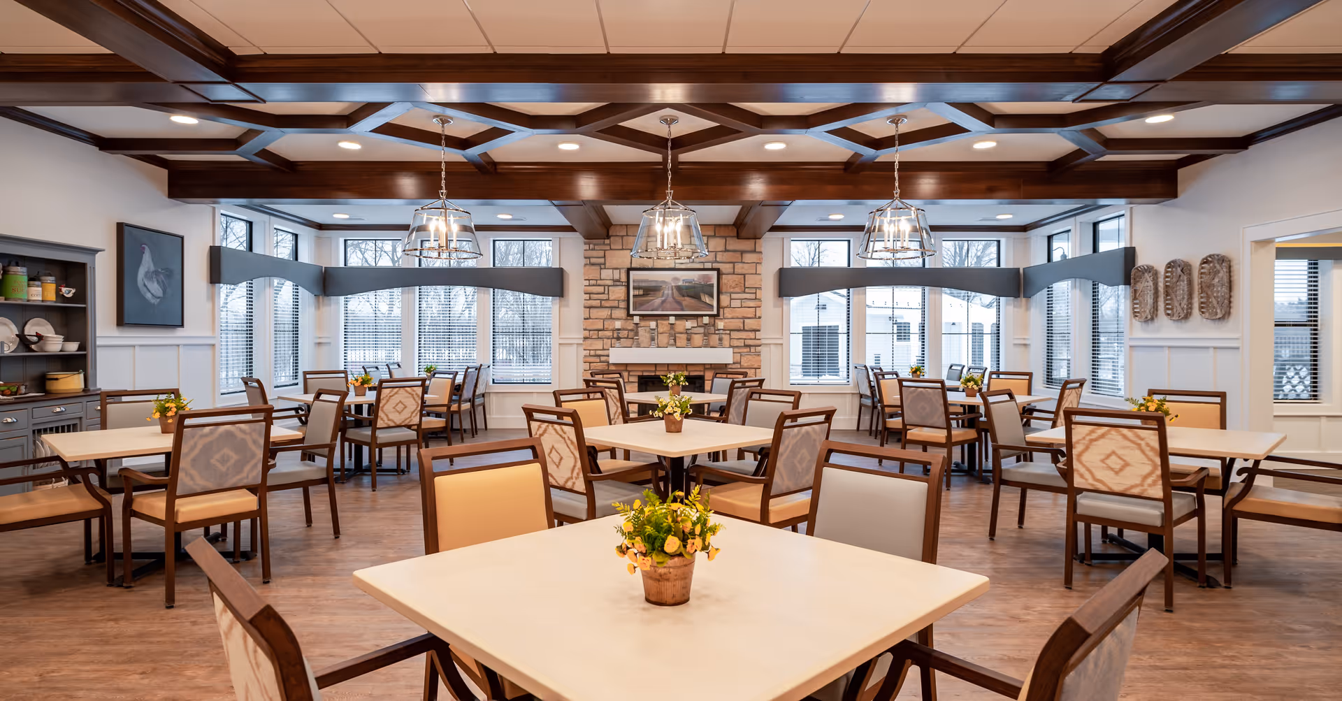 A spacious and well-lit dining room in a senior living facility with multiple square tables and cushioned chairs arranged neatly. Each table has a small flower arrangement. The room features large windows with blinds, a stone fireplace with a framed picture above it, wooden ceiling beams, and modern pendant lights hanging from the ceiling.