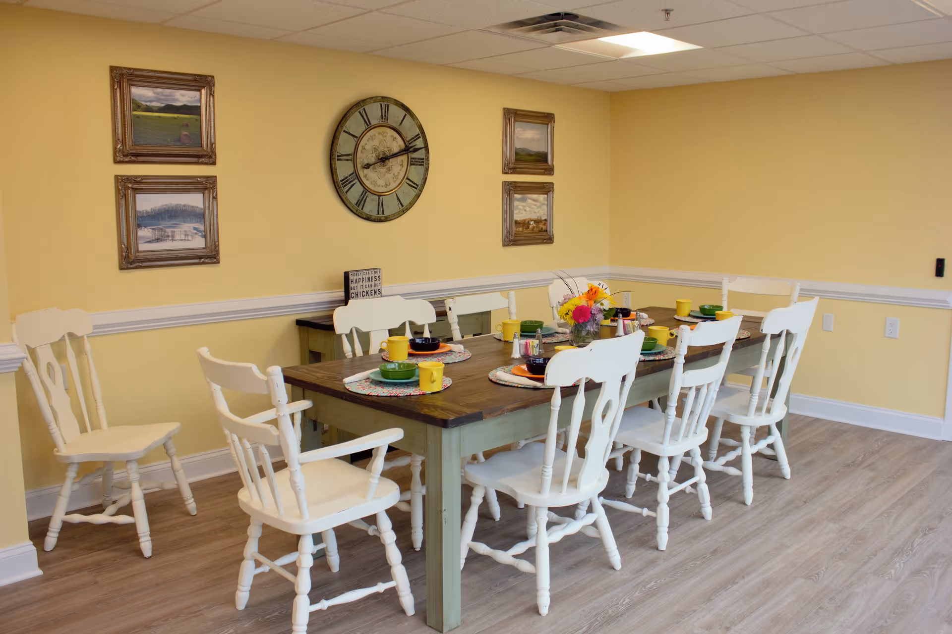 A dining room with a wooden table set for six people. The table has colorful bowls, plates, and mugs, along with a vase of flowers in the center. The room has yellow walls with white trim, a large round clock, and four framed landscape paintings. The floor is light wood, and there are white wooden chairs around the table.