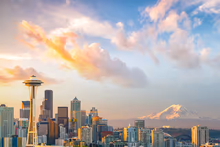 View of the Seattle skyline at sunset with the iconic Space Needle prominently visible on the left and Mount Rainier in the background under a partly cloudy sky.