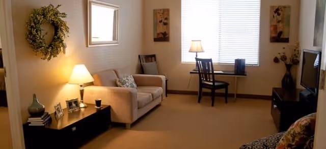 A cozy living room with a beige sofa, a side table with a lamp and decorative items, a chair and desk near a window with blinds, a TV on a stand, and wall decorations including a wreath and framed pictures.