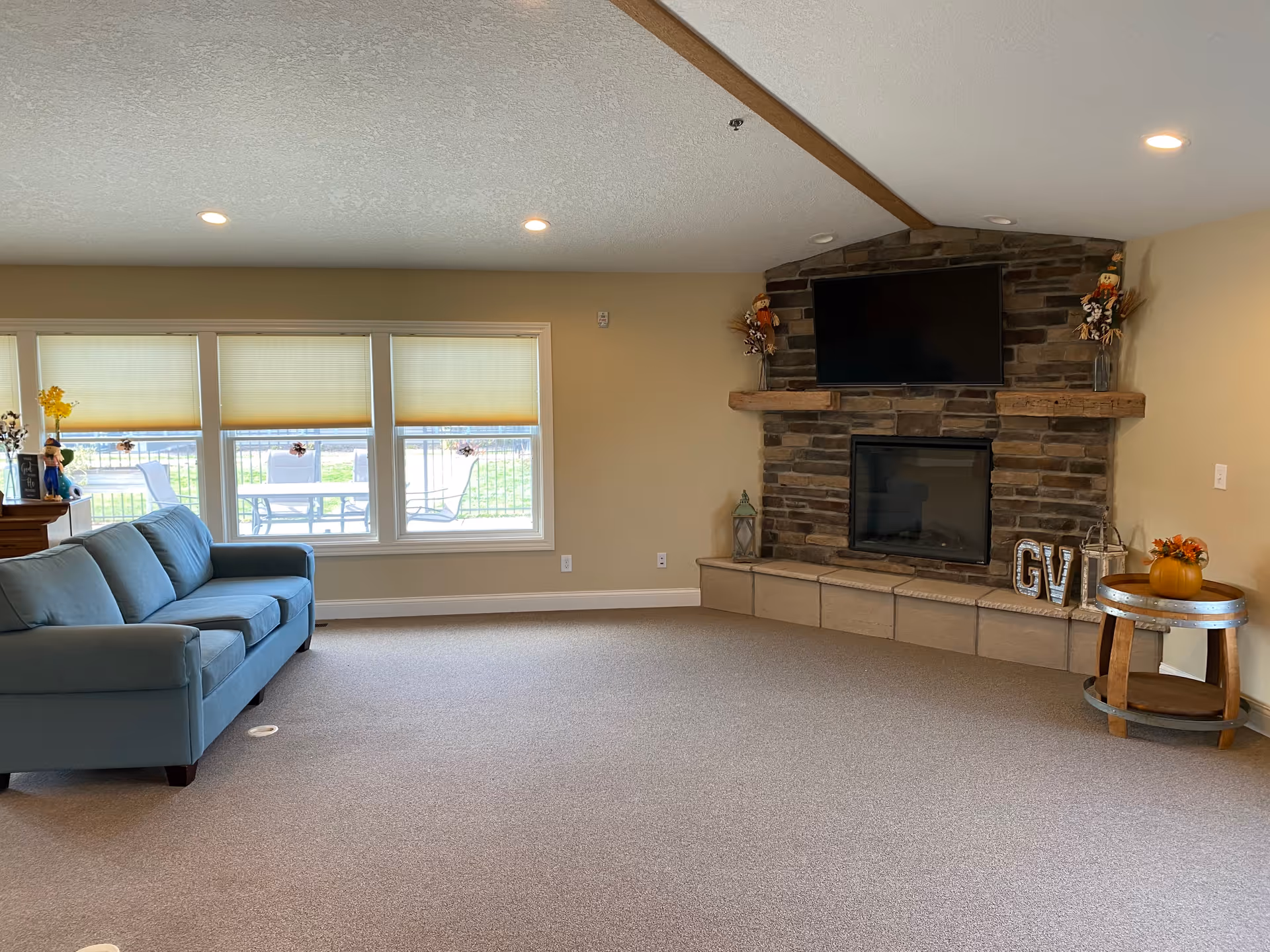 A spacious living room with a light blue sofa on the left, large windows with beige blinds behind it, and a stone fireplace with a mounted flat-screen TV above it on the right. The fireplace mantel is decorated with small scarecrow figures and other fall-themed decor. A round wooden side table with a small pumpkin and flower arrangement is placed near the fireplace. The room has beige walls and carpeted flooring.