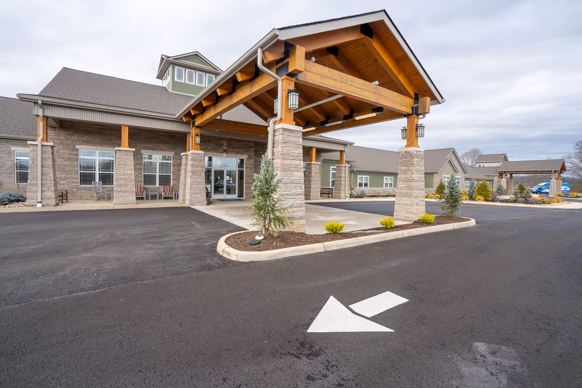 Exterior view of Princeton Transitional Care facility showing the main entrance with a covered drop-off area supported by stone pillars and wooden beams. The building has a combination of stone and siding with multiple windows and a paved driveway with a white directional arrow painted on the asphalt.