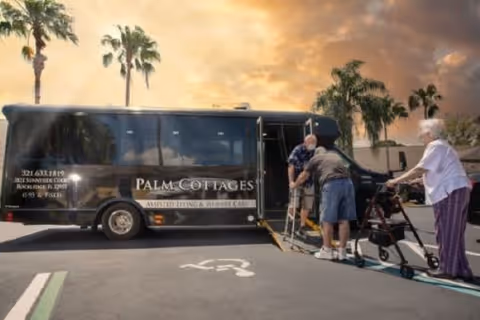 A Palm Cottages of Rockledge shuttle bus parked in a lot with a sunset sky in the background. Two elderly individuals are assisting a third person using a walker to board the bus via a ramp. Palm trees are visible around the parking area.
