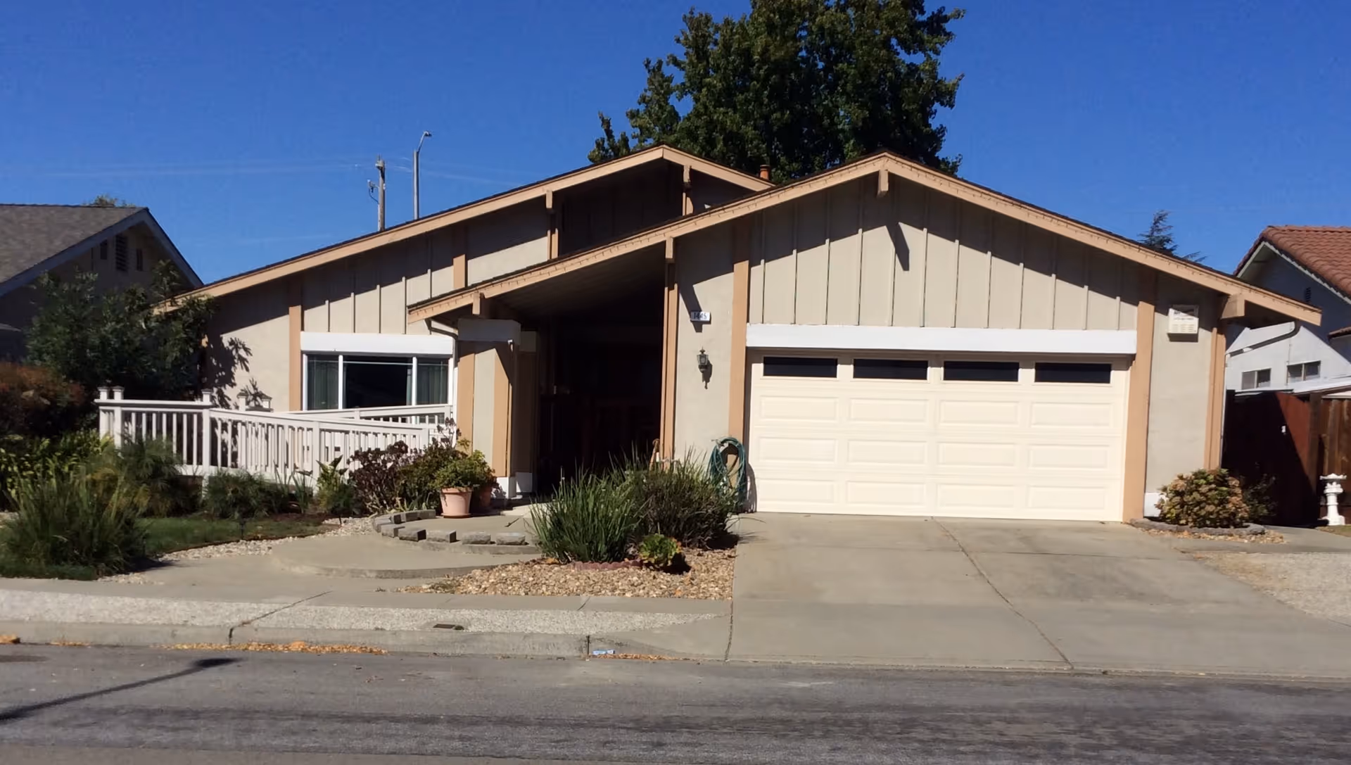 Front exterior view of a single-story residential building with a beige and brown color scheme, a white garage door, a driveway, and a small garden with plants and shrubs. The sky is clear and blue.
