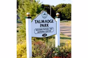 White sign with black text reading 'Talmadge Park Rehabilitation and Nursing Center 38' mounted on two white posts with gold-colored finials, surrounded by greenery and trees in the background.