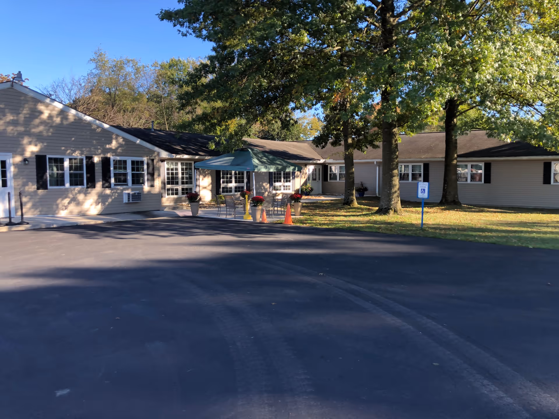 Single-story facility front with a paved driveway, trees, outdoor seating under a canopy, and a handicapped parking sign.