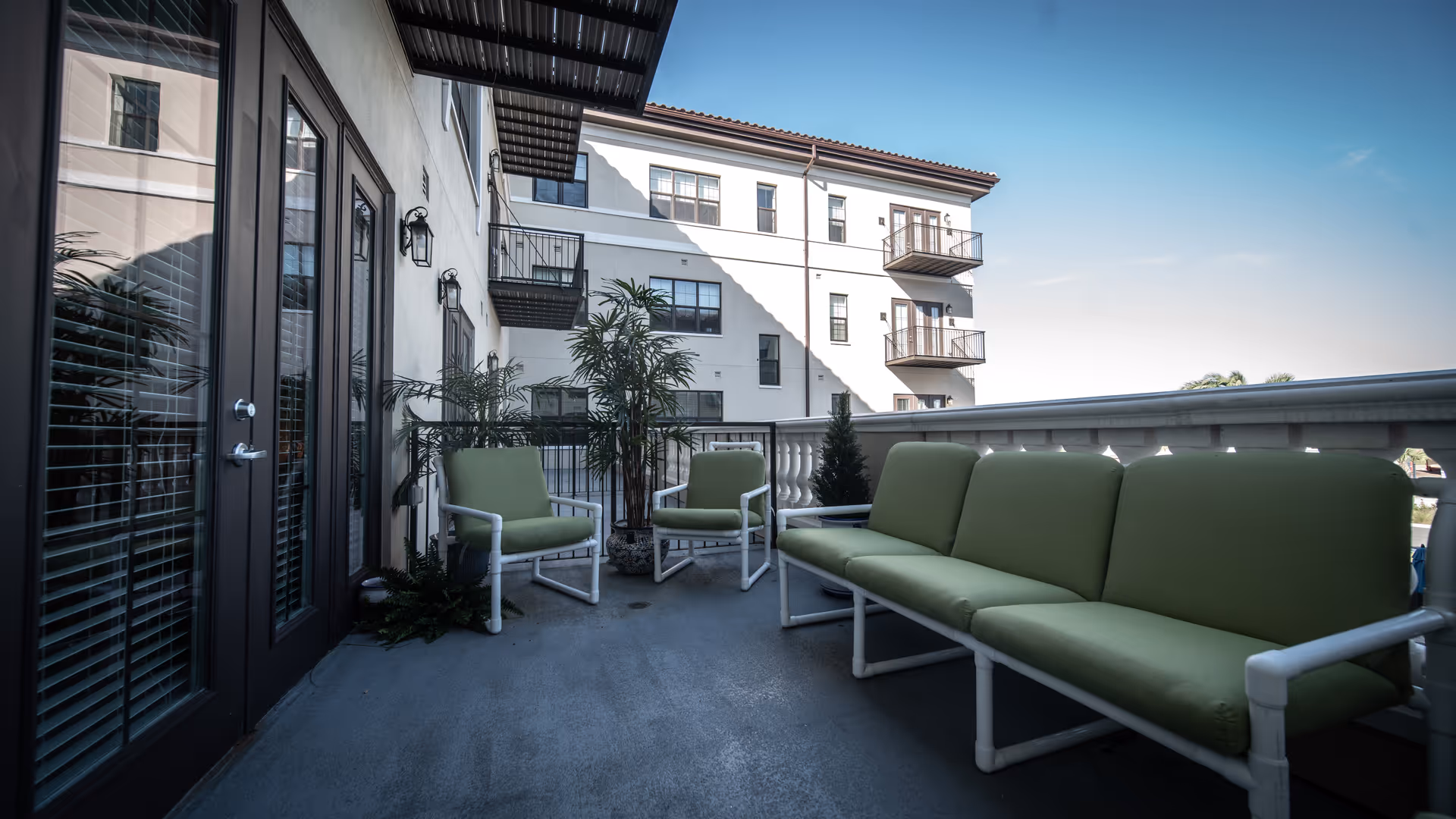 Outdoor balcony area with green cushioned seating including a long sofa and two chairs, potted plants, and a view of a multi-story building with balconies under a clear blue sky.