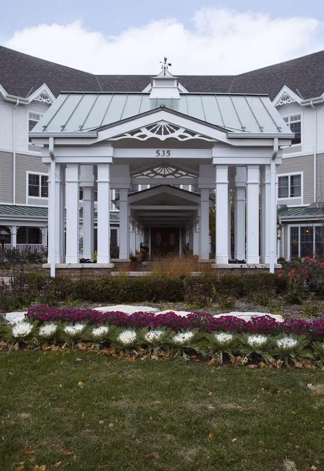 Front exterior view of a senior living facility with a covered entrance supported by white columns, a green metal roof, and the number 535 displayed above the entrance. There are flower beds with purple and white flowers in front of the building and a well-maintained lawn.