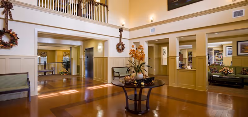 Spacious and well-lit interior common area with polished wooden floors, a round table with a floral arrangement in the center, green cushioned benches along the walls, decorative wreaths hanging on the walls, and an upper balcony overlooking the space.