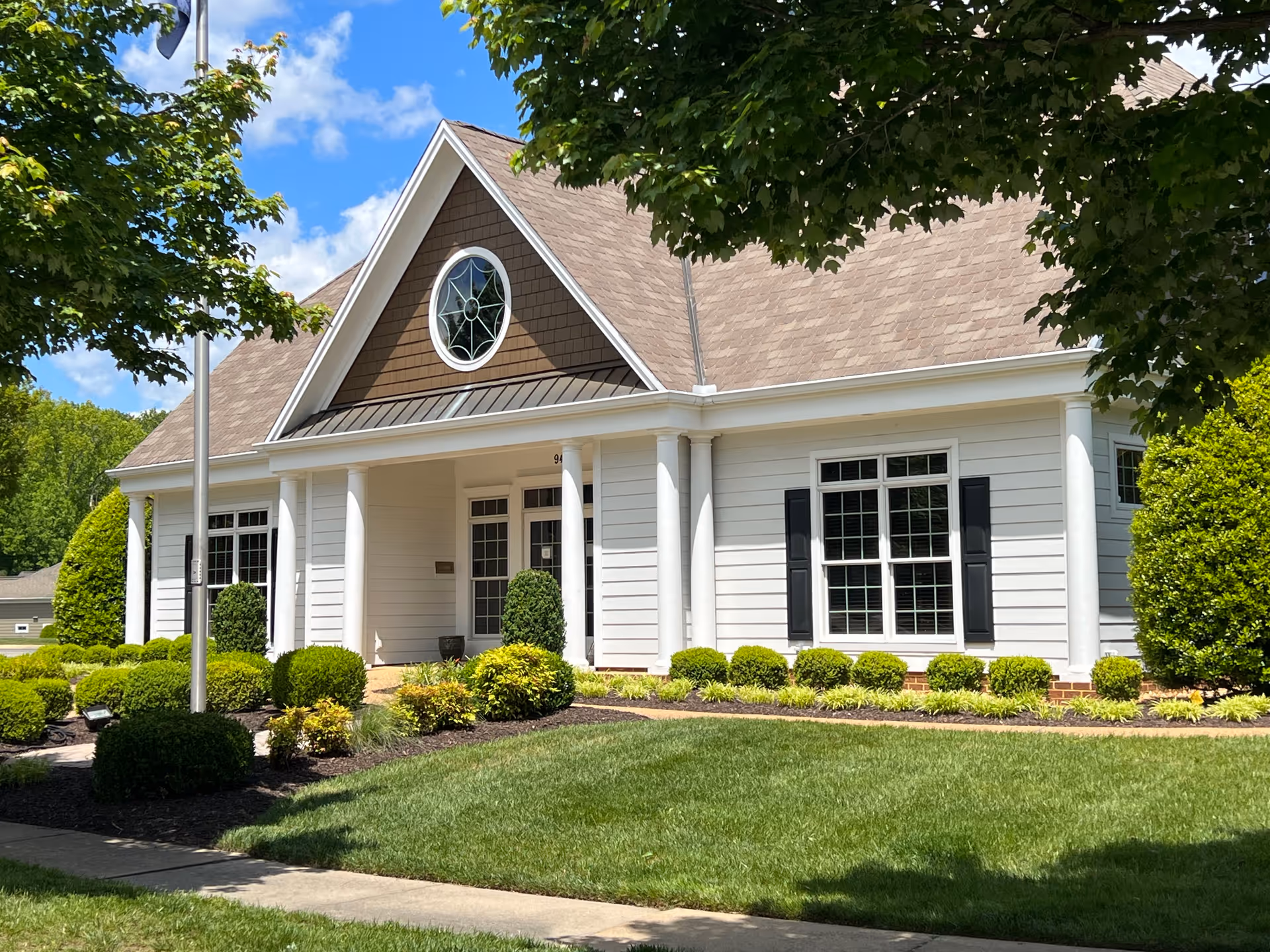 Front exterior of a white building with columns, large windows, a circular gable window, and manicured landscaping.