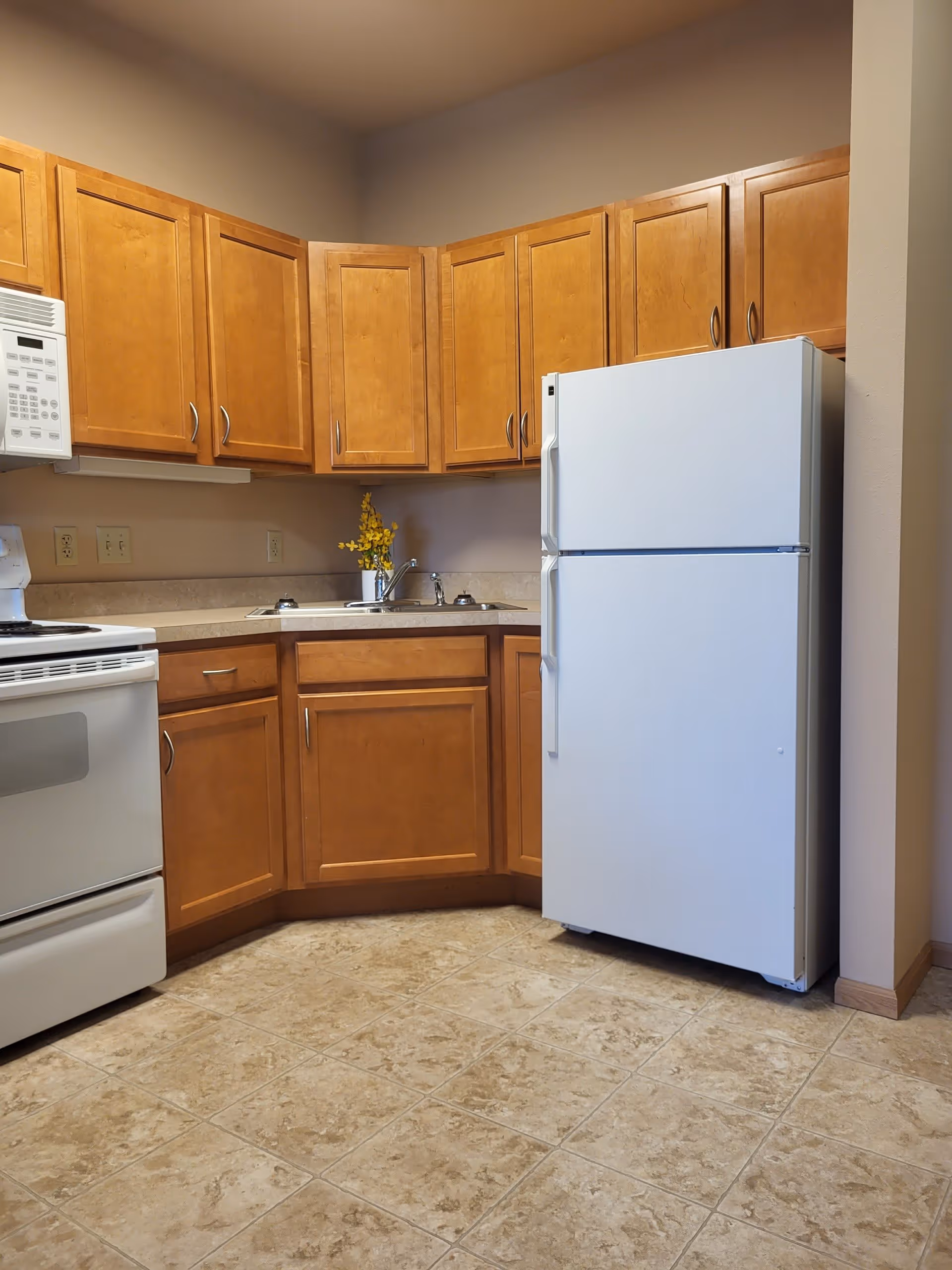 A kitchen area with wooden cabinets, a white refrigerator, a white stove with an oven, a white microwave, a countertop with a sink, and a small vase with yellow flowers. The floor is tiled with a beige pattern.