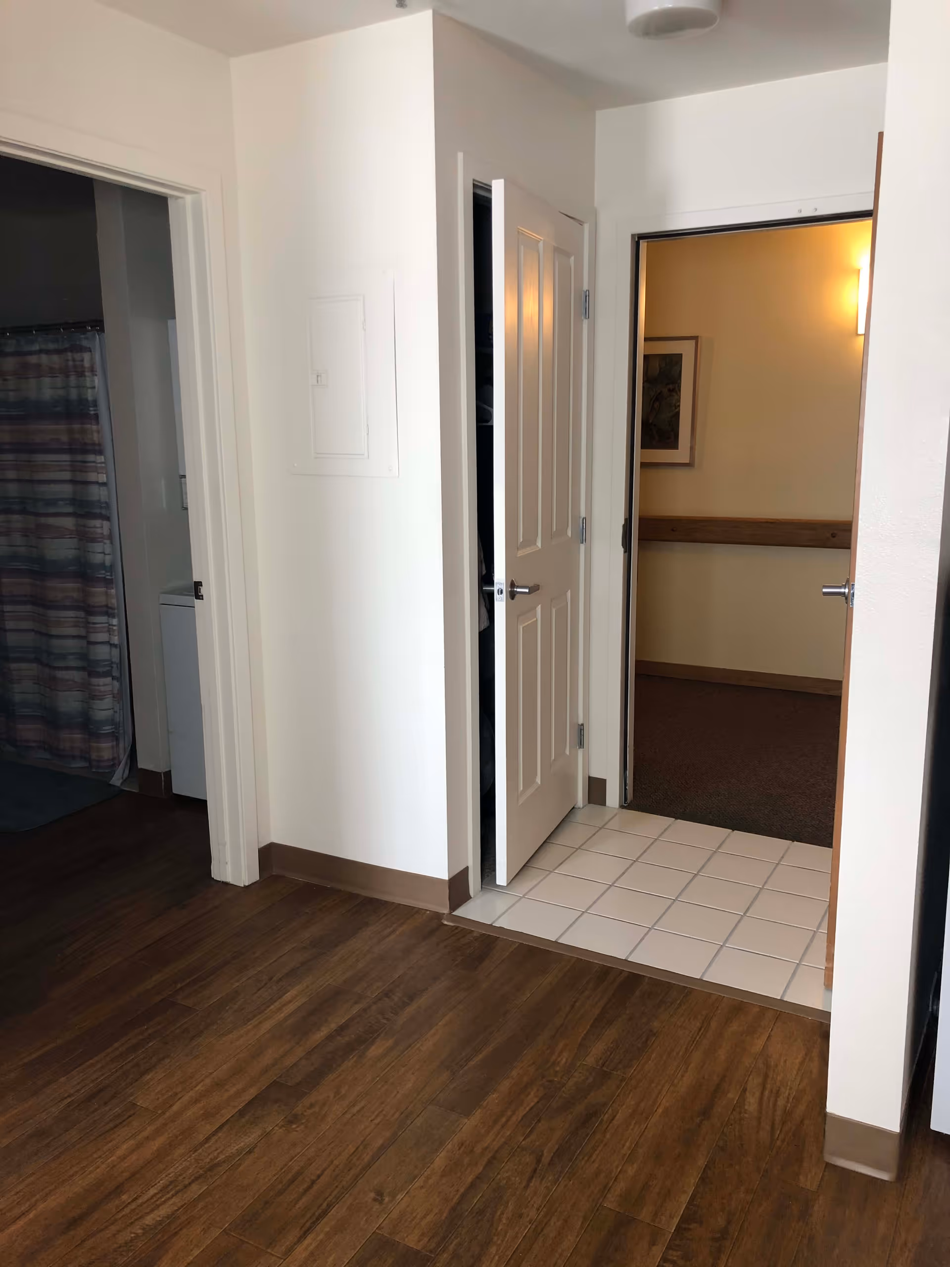 Interior view of a hallway in a senior living facility with wooden flooring and white walls. There are three doorways visible: one leading to a bathroom with a shower curtain, one with a partially open door revealing a closet, and one leading to a carpeted room with a framed picture and wall light.