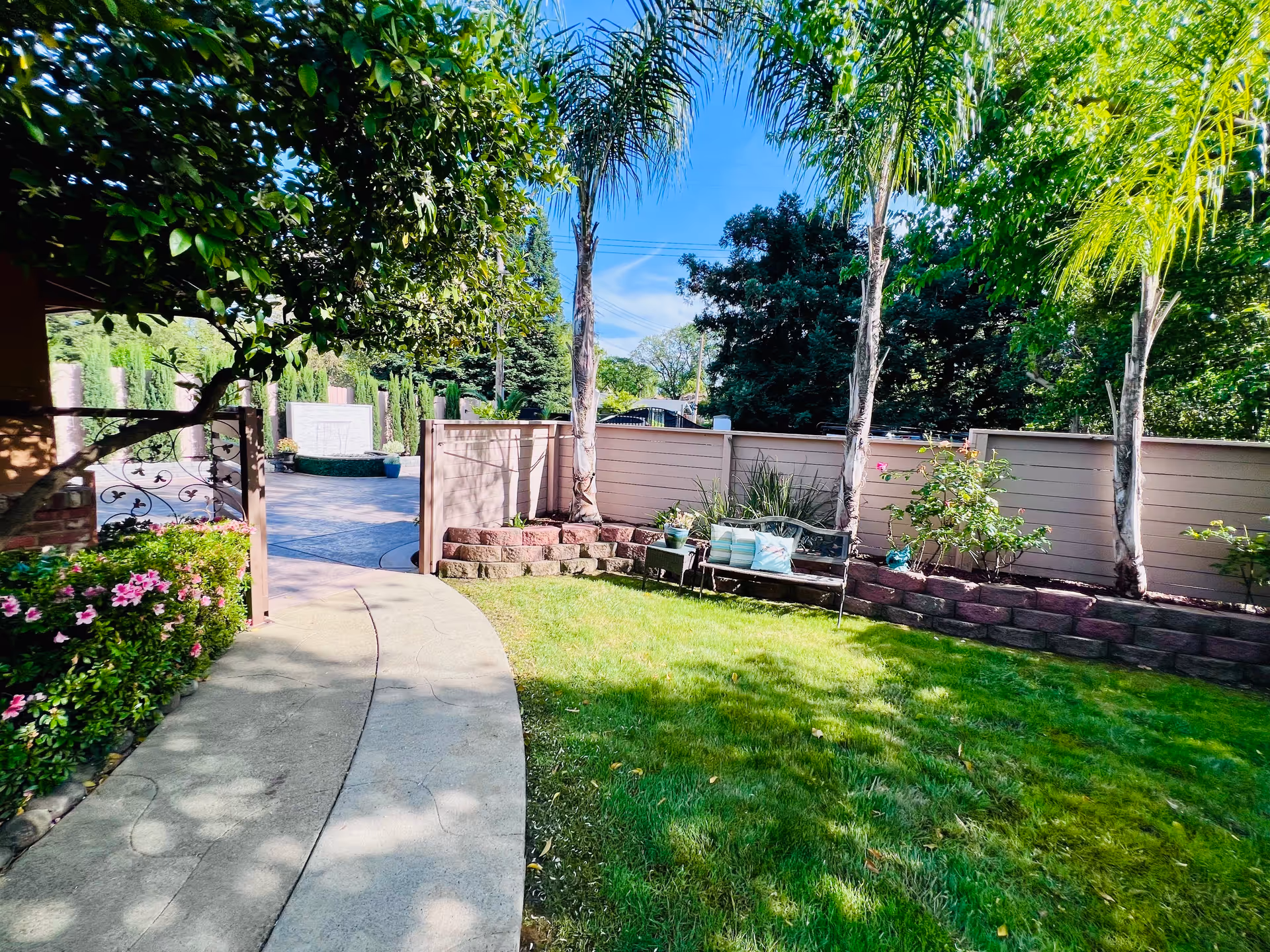 A sunny outdoor garden area with a curved concrete pathway, green grass, palm trees, flowering plants, and a bench with cushions against a wooden fence.