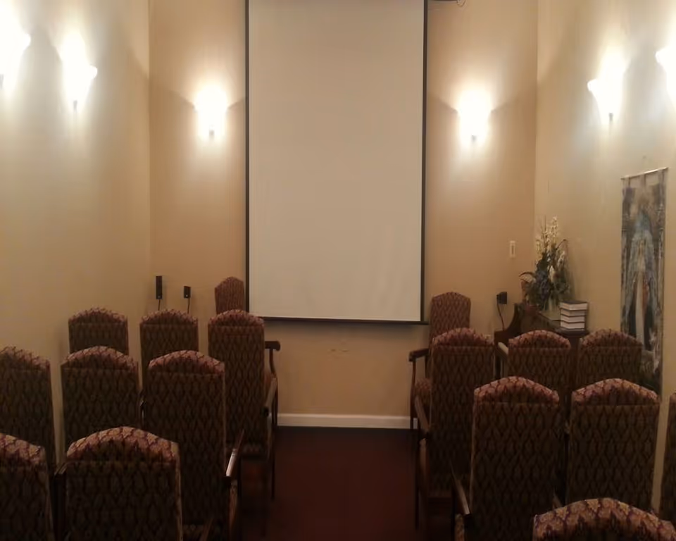 Rows of patterned upholstered chairs facing a blank projection screen in a small meeting/screening room with wall sconces and a floral arrangement.