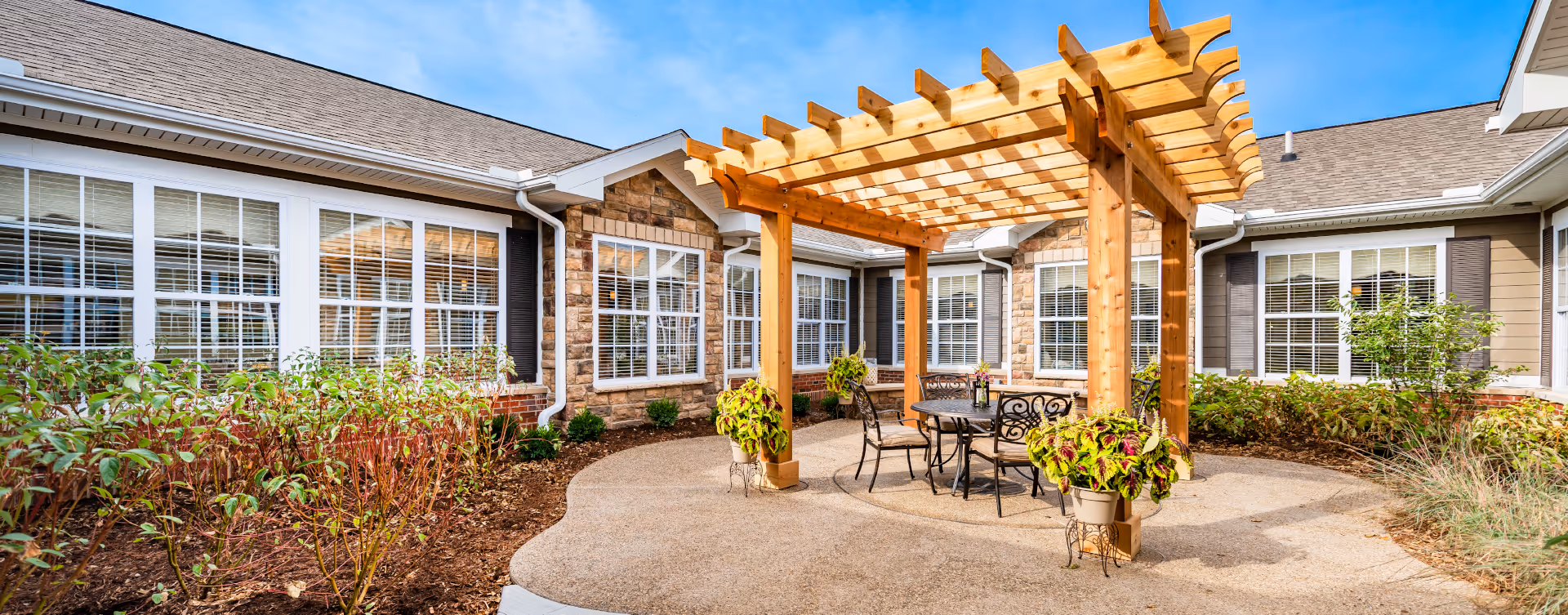 Outdoor patio area at Bickford of Shelby Township featuring a wooden pergola with a round metal table and four chairs underneath. The patio is surrounded by plants and bushes, with the building's windows and stone exterior visible in the background under a clear blue sky.