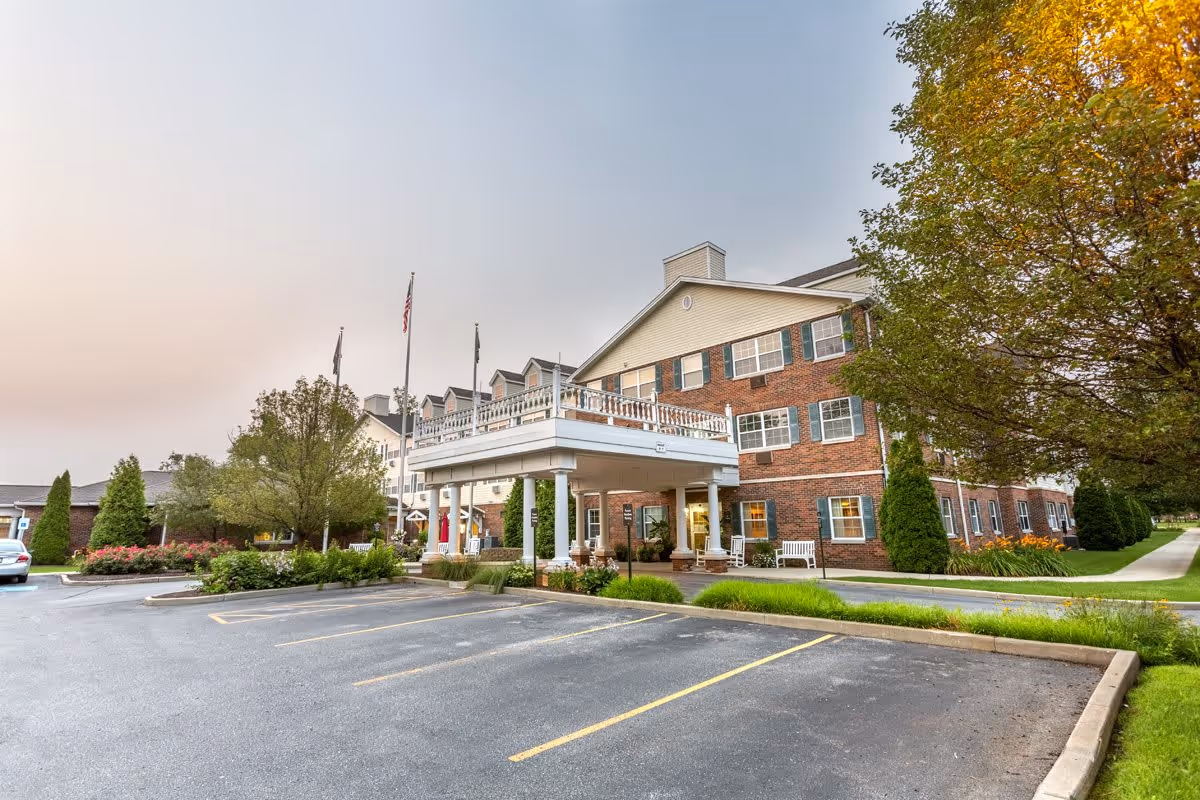 Exterior view of a multi-story senior living facility with a brick facade and white trim. The entrance features a covered drop-off area with white railings and columns. There are several flagpoles with flags near the entrance. The surrounding area includes a parking lot, trees, shrubs, and a well-maintained lawn.