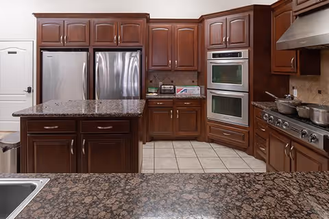 A spacious kitchen with dark wooden cabinets, granite countertops, a large kitchen island, stainless steel refrigerator, double wall ovens, and a gas stove with pots on it. The floor is tiled and there is a white door with a black handle on the left side.