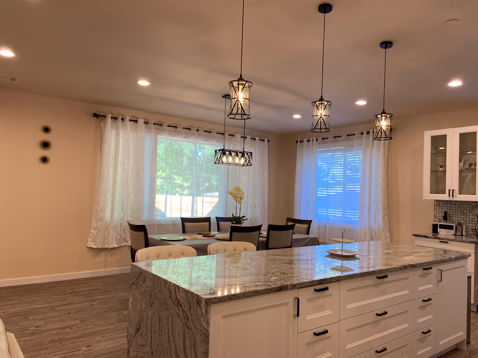 A bright and modern dining area and kitchen with a large marble island in the foreground. The dining table is set with chairs and a centerpiece, positioned near two large windows with white curtains. Three pendant lights hang from the ceiling above the island and dining table. The kitchen cabinets are white with black handles, and there is a tiled backsplash visible.