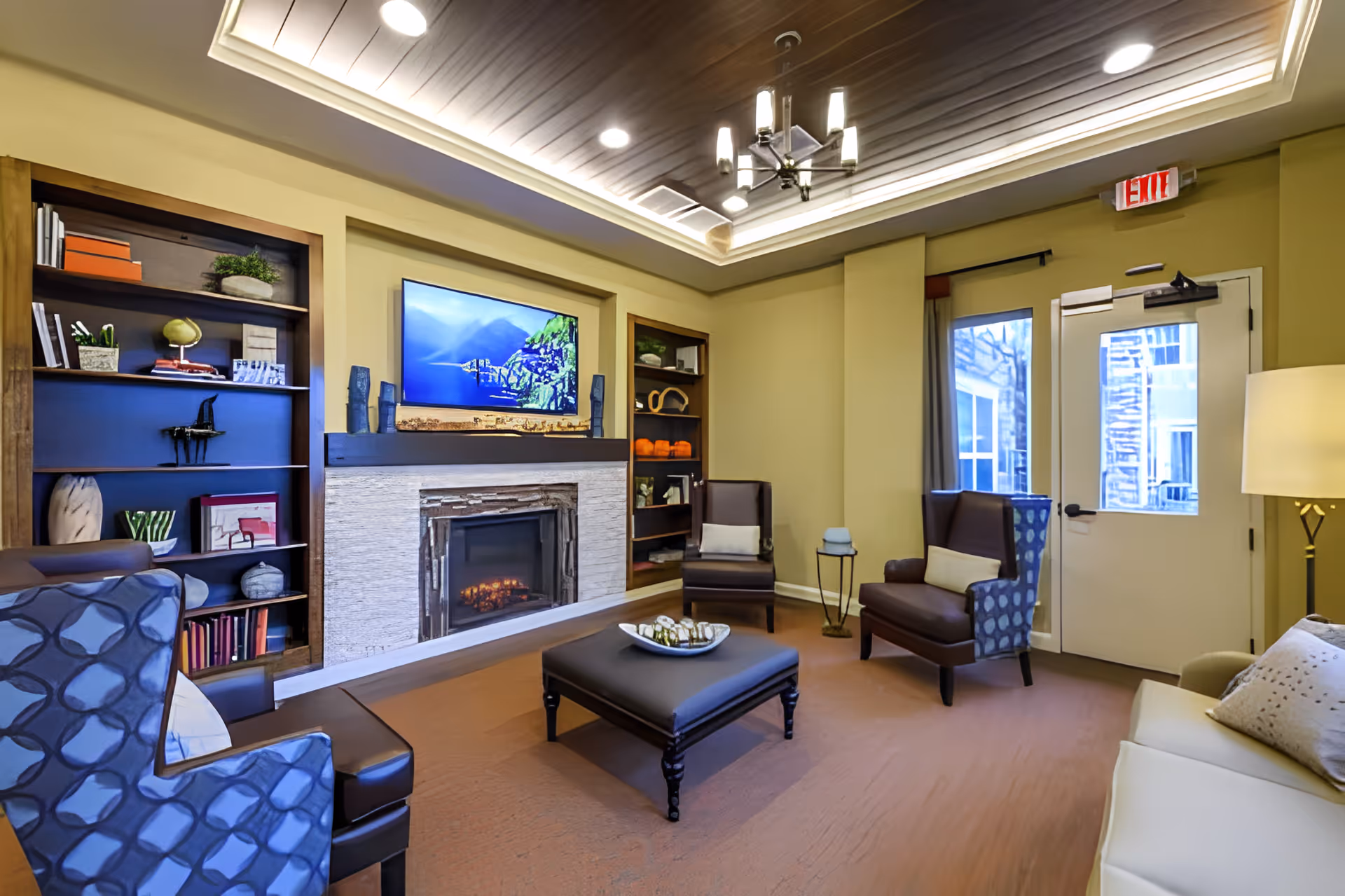 A bright common living room with a fireplace, wall-mounted TV, bookshelves, and several upholstered chairs and a sofa.