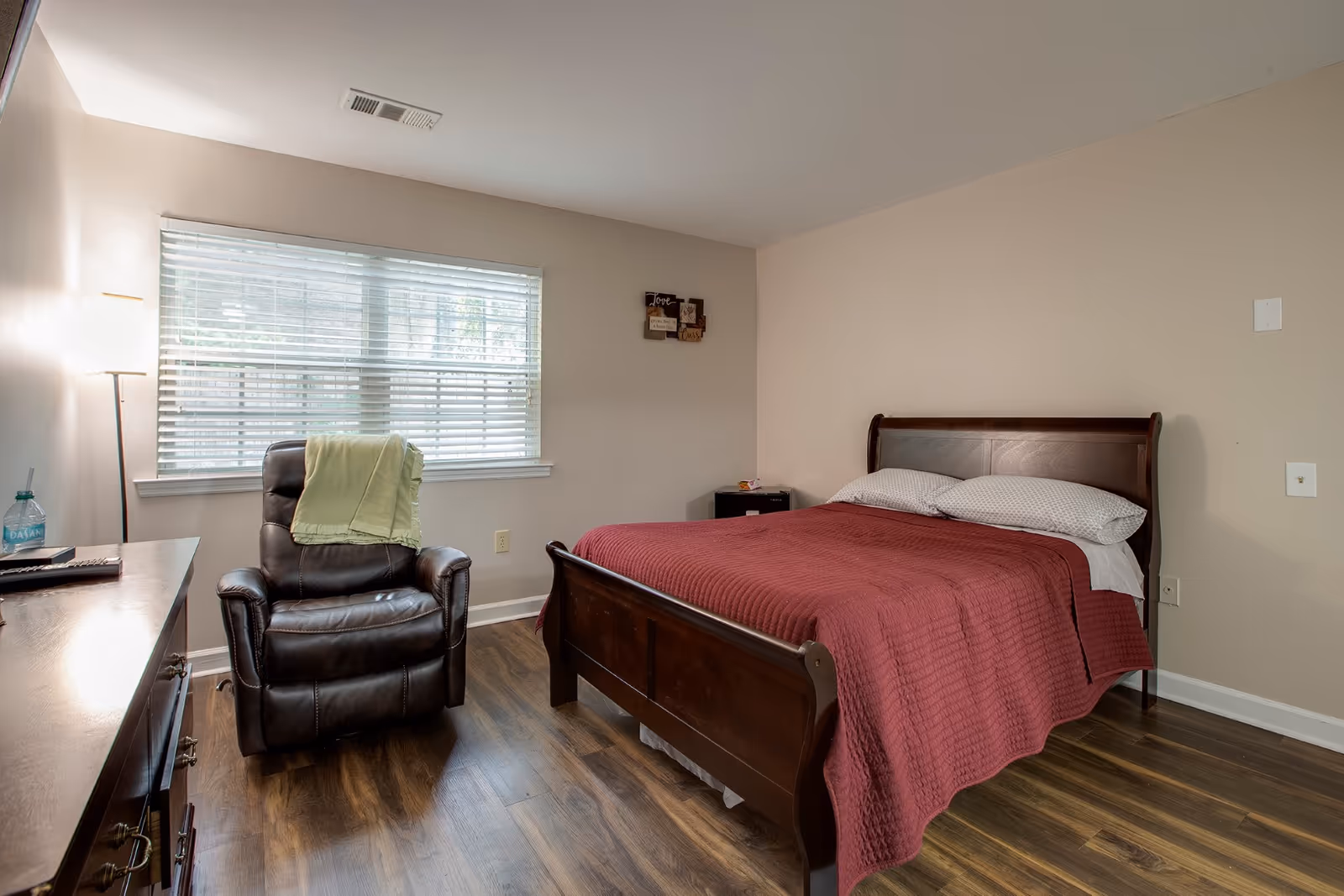 A tidy bedroom with a wooden bed covered by a red quilt, a leather recliner by a window, and a dresser.