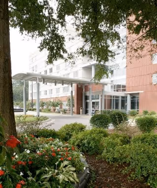 Front entrance of a multi-story senior living building with a covered drop-off and landscaped flower beds in the foreground.
