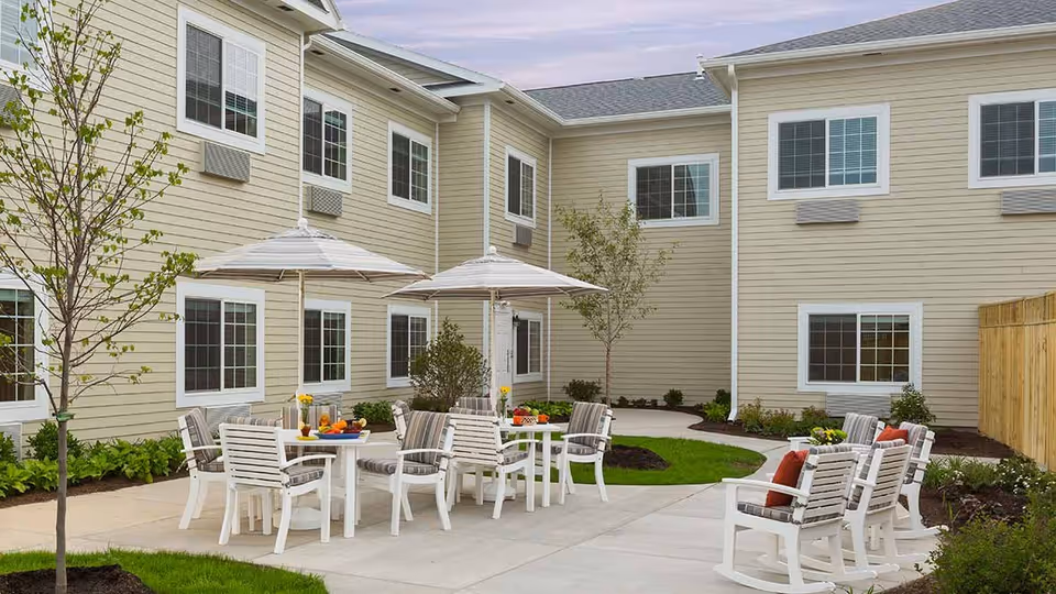 Outdoor patio area at The Landing Of Brighton with white tables and chairs, some with striped cushions and umbrellas, surrounded by a beige two-story building with multiple windows and small trees and plants along the walkway.