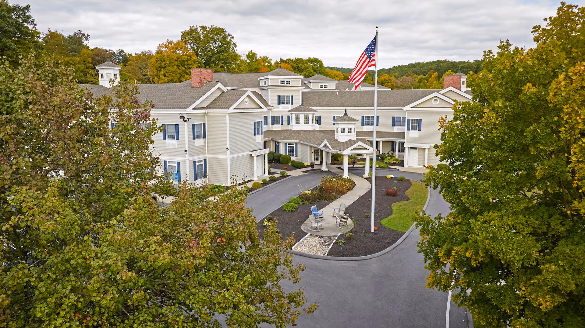 Front exterior of a multi-story senior living building with a circular driveway, flagpole, and surrounding trees.