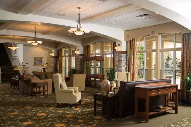 Sunlit communal lounge with upholstered armchairs, sofas, side tables, decorative lamps, and large windows overlooking the grounds.