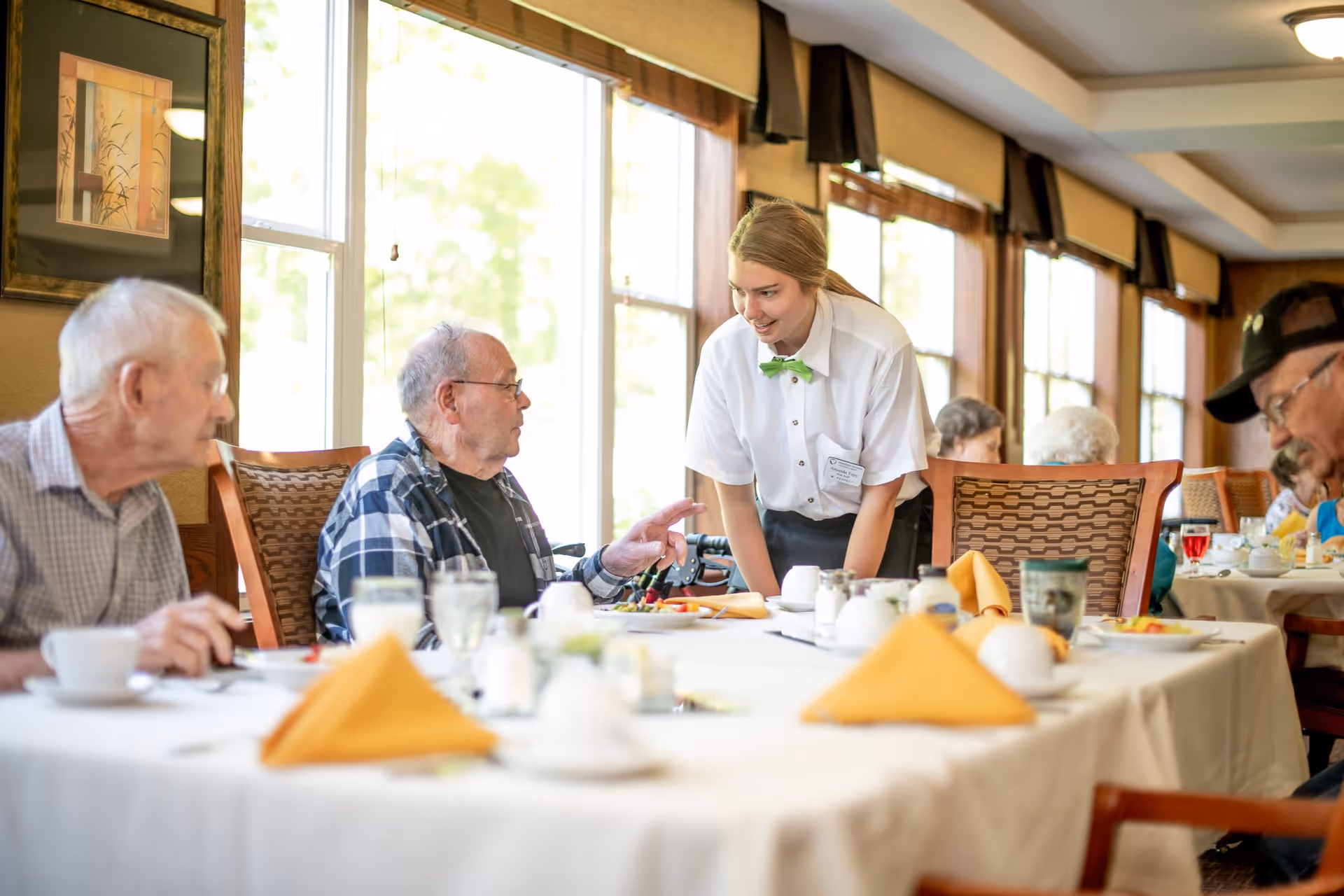 A server leans forward speaking with elderly residents seated at a long, well-set dining table in a bright dining room.