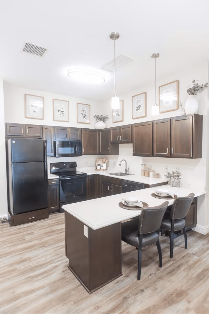 A modern kitchen with dark wooden cabinets, black appliances including a refrigerator, stove, and microwave, and a white countertop island with two place settings and two black chairs. The kitchen has light wood flooring, pendant lights hanging from the ceiling, and framed botanical prints on the walls above the cabinets.