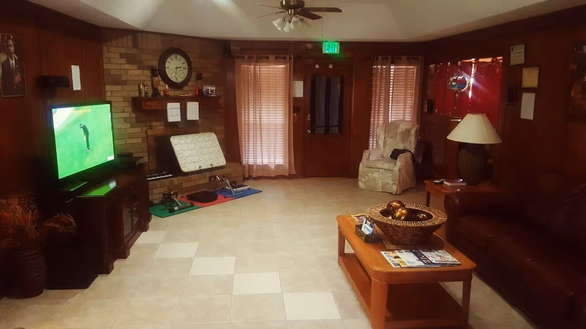 A cozy living room with a tiled floor, a brown leather sofa, a wooden coffee table with a basket and magazines, a floral armchair, a table lamp, and a television showing a golf game. The room has wood-paneled walls, a brick fireplace with a clock above it, and two windows with blinds and curtains. There is an exit door with an illuminated green exit sign above it.