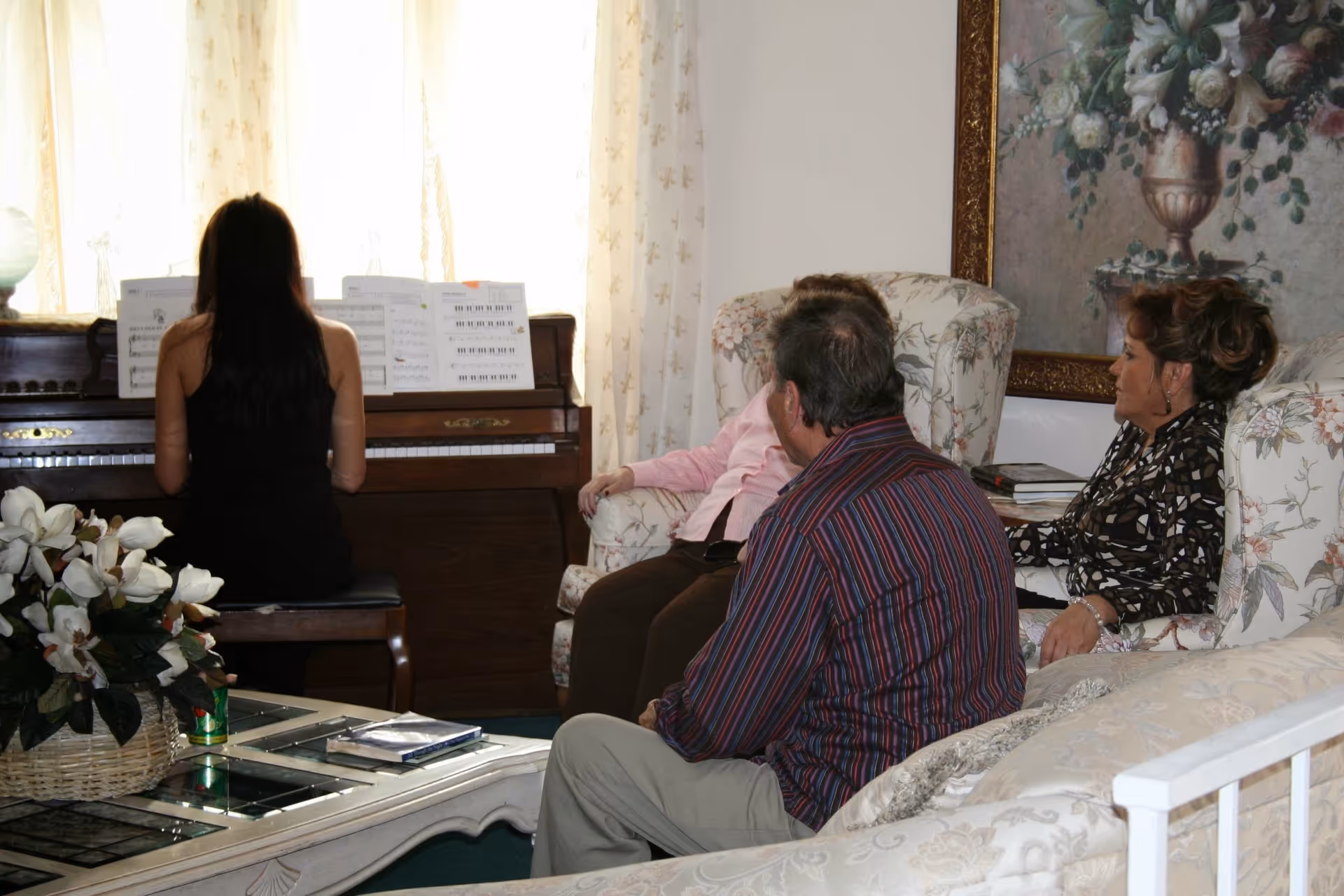 A woman playing piano while three elderly people sit on floral upholstered chairs and a sofa in a living room with a large floral painting on the wall and a coffee table with a flower arrangement.