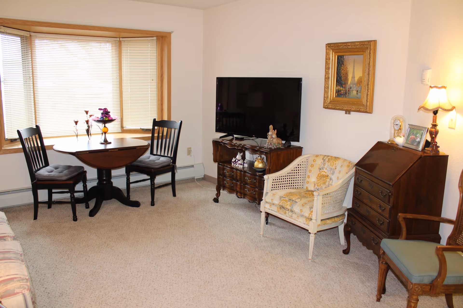 Cozy living room with a bay-window table and chairs, a TV on a wooden cabinet, and antique-style chairs and furniture.