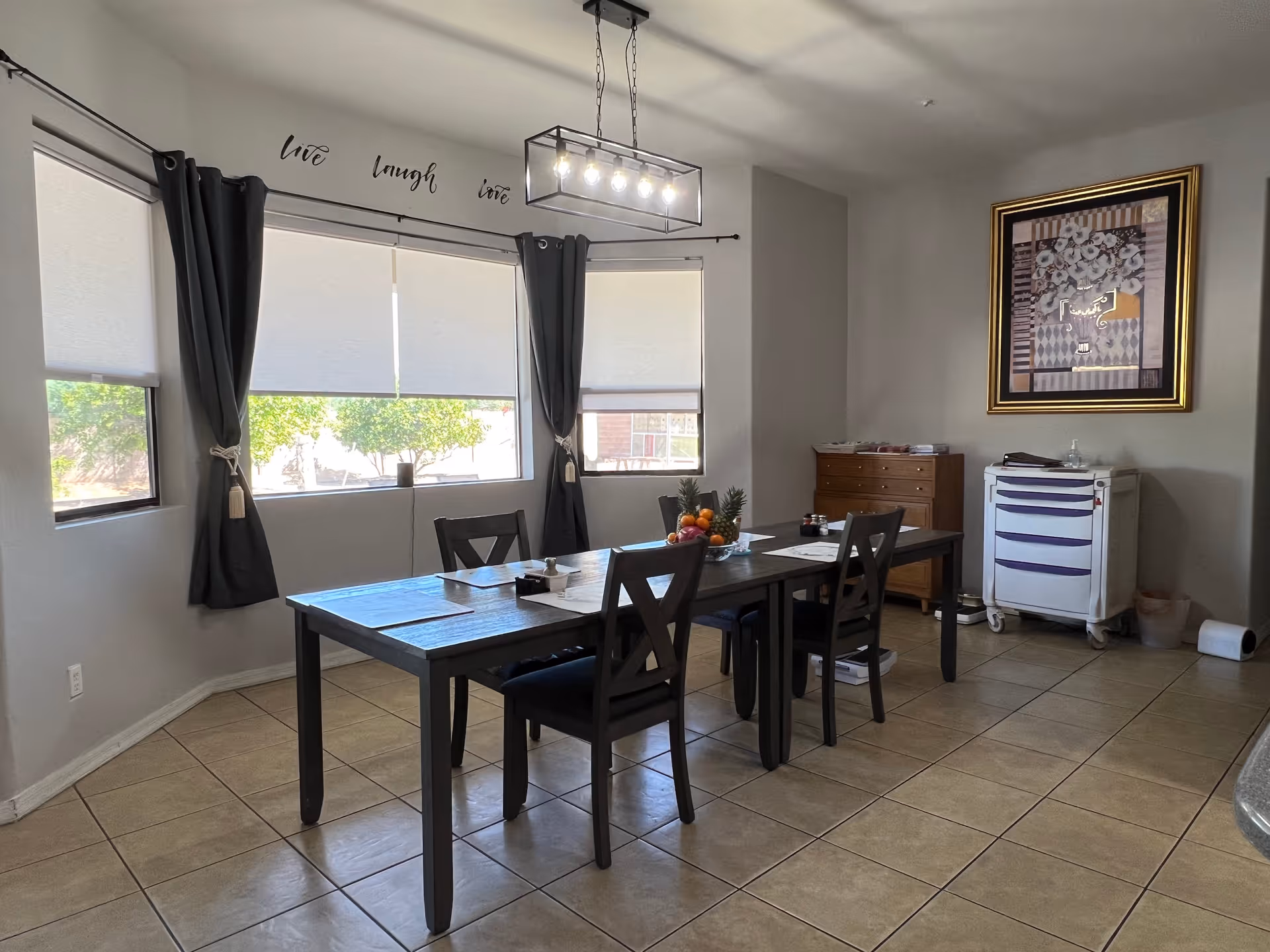 A dining room with a rectangular wooden table and four chairs. The table has placemats, a bowl of fruit, and some small items on it. The room has large windows with white roller blinds and dark curtains. Above the windows, the words 'live laugh love' are displayed on the wall. A modern rectangular light fixture hangs above the table. There is a framed artwork on the wall and a small cabinet and a medical cart in the corner. The floor is tiled.