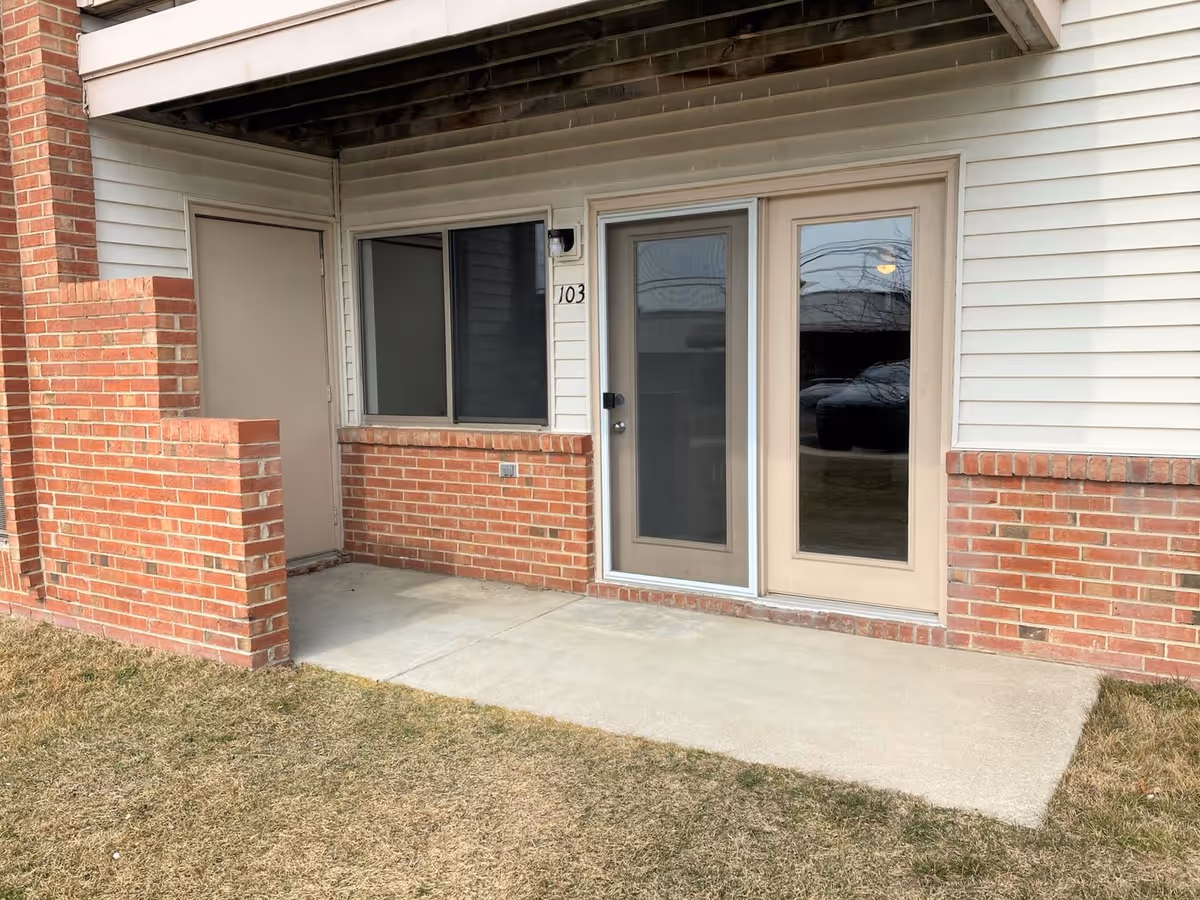 Exterior view of a senior living facility unit with a small concrete patio, brick half-wall, sliding glass door, a window, and a solid door. The unit number 103 is visible next to the sliding door.
