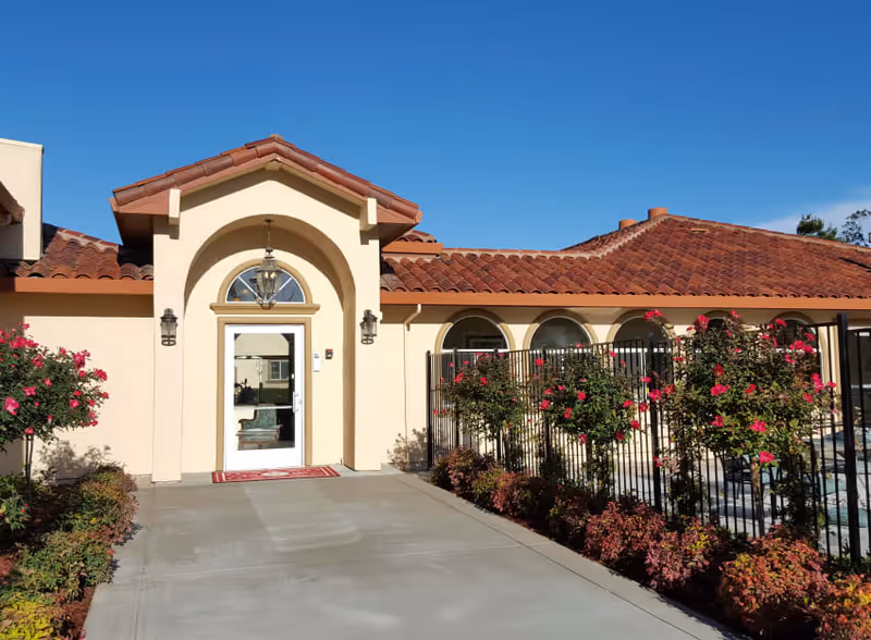 Exterior view of a single-story building with beige walls and a red tiled roof under a clear blue sky. The entrance features an arched doorway with a glass door and a hanging lantern light. There are flowering bushes and a black metal fence along the right side of the walkway leading to the entrance.