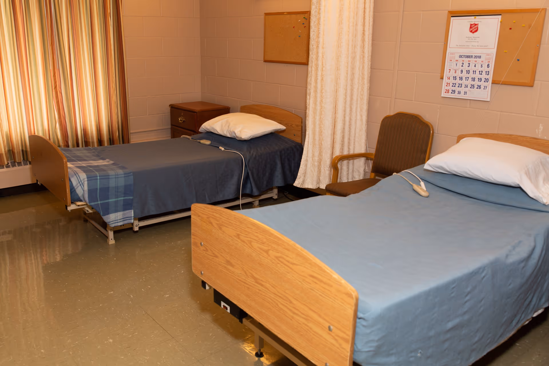 A room with two single hospital-style beds, each with a pillow and blue bedding. There is a wooden nightstand between the beds, a brown chair, a curtain divider, and a calendar on the wall showing October 2018. The room has beige walls and a window with striped curtains.