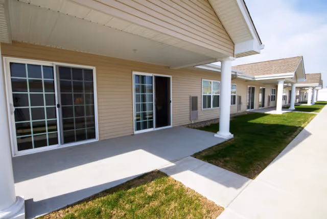 Exterior view of a single-story building with beige siding, white columns, sliding glass doors, and windows. There is a concrete walkway and small patches of grass along the building under a clear sky.