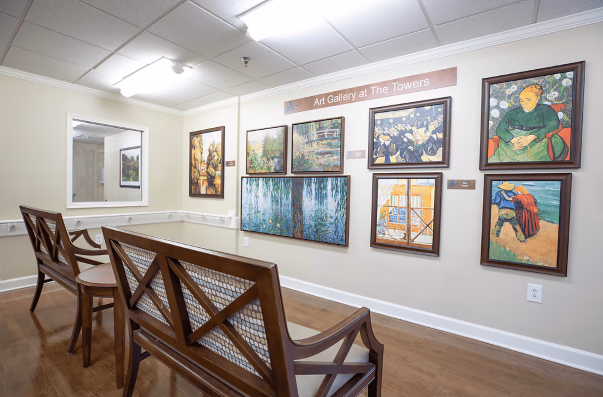 Interior view of an art gallery area in a senior living facility with wooden benches facing a wall displaying several framed paintings. The wall has a sign reading 'Art Gallery at The Towers'. The floor is wooden, and the ceiling has recessed lighting.
