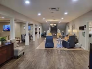 A spacious and well-lit living area in a senior living facility with wooden flooring, comfortable armchairs, a small round table, and a staircase in the background. The room features neutral-colored walls, ceiling lights, and a television mounted on the wall to the left.