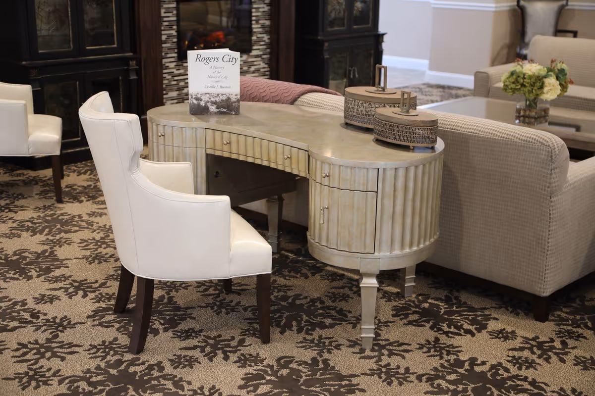A cozy interior seating area featuring a curved beige desk with drawers, a white upholstered chair, and beige sofas. On the desk is a book titled 'Rogers City: A History of the Nautical City' and two decorative round containers. The room has a patterned carpet and a fireplace in the background.