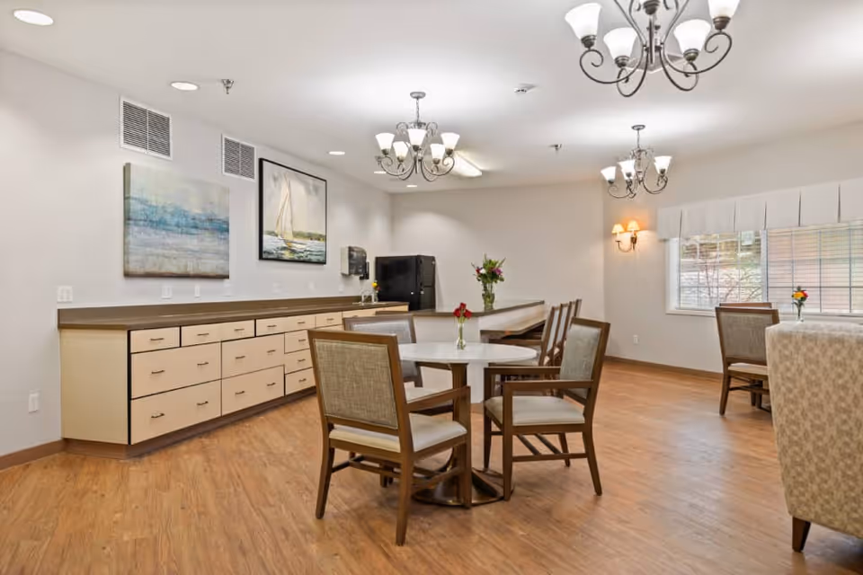 A bright and clean dining area in Aspen Ridge Memory Care featuring wooden floors, round tables with chairs, a long counter with multiple drawers, two framed paintings on the wall, a coffee machine, and ceiling chandeliers providing warm lighting.